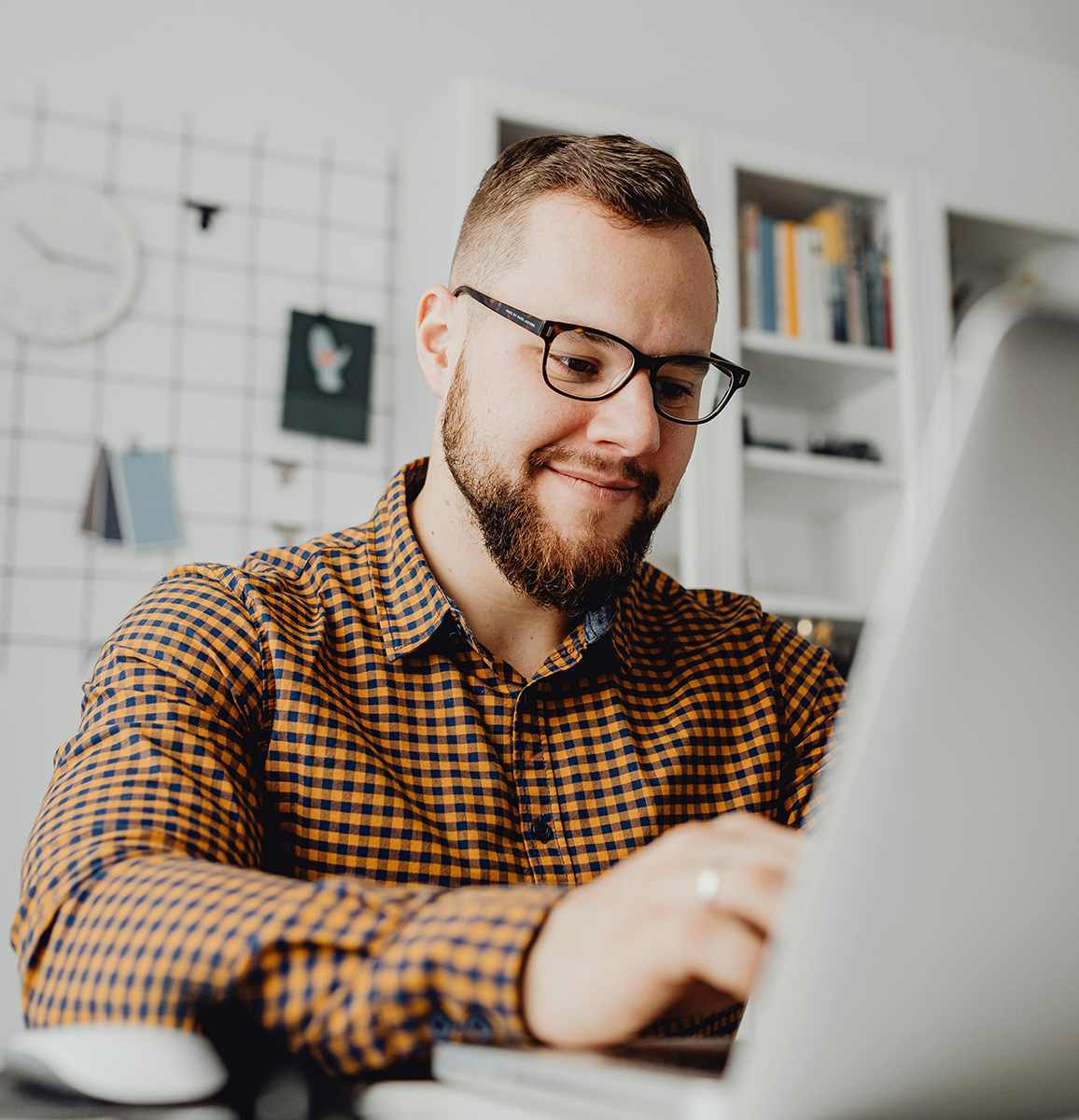 Bearded man with glasses smiling as he types at a laptop in a brightly lit white room with a book shelf in the background.